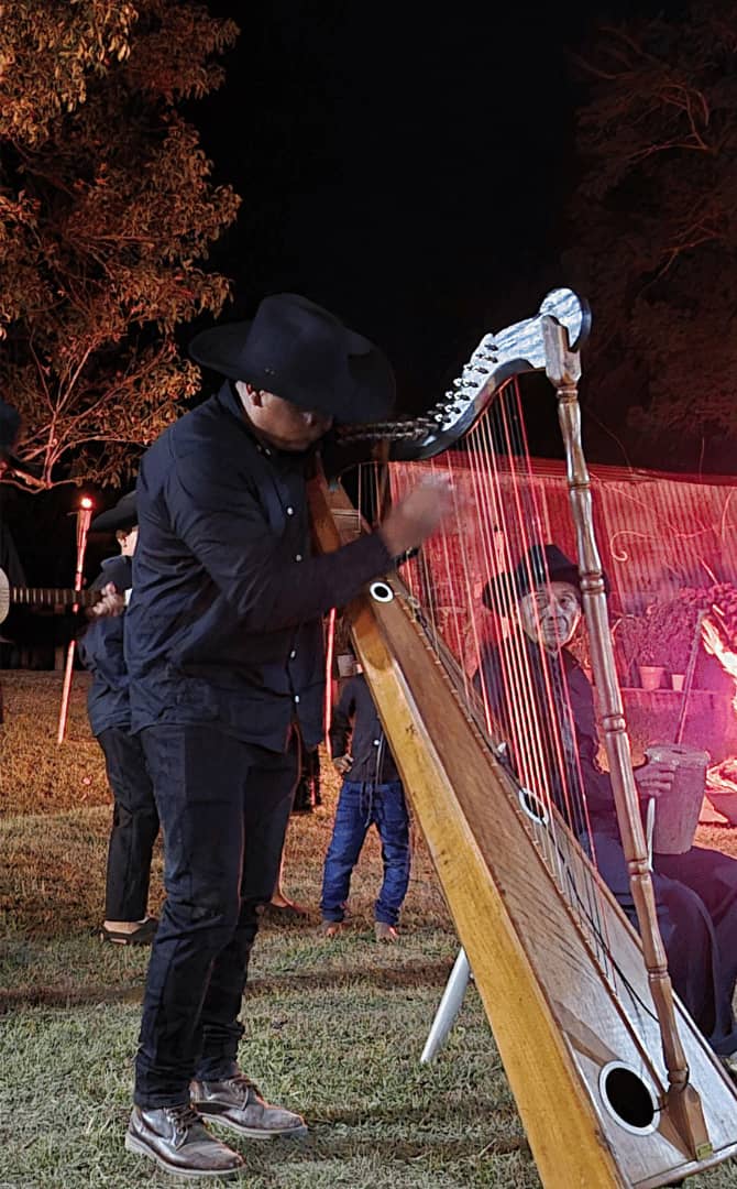 Perfil detallado de un músico llanero tocando un arpa llanera de madera durante una presentación nocturna en el campo con otros artistas.