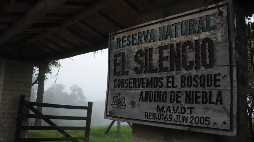 Plano detalle en ángulo contrapicado del letrero rústico de madera de la Reserva Natural El Silencio, donde se lee grabado 'Reserva Natural El Silencio - Conservemos el bosque andino de niebla'. El fondo muestra una densa neblina blanca y la silueta borrosa de árboles de montaña.