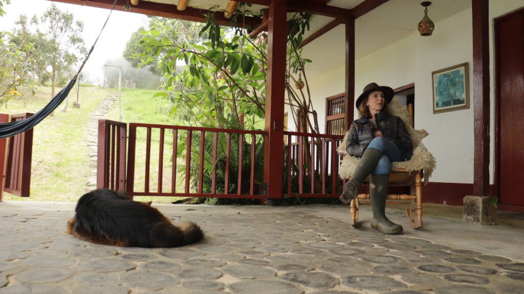 Fotografía de mujer sentada en una mecedora de madera con una manta de lana en el corredor rústico de la casa principal de la Reserva Natural El Silencio. Viste botas de caucho, sombrero de ala ancha y chaqueta, mientras conversa en un entorno rodeado de vegetación andina y neblina.