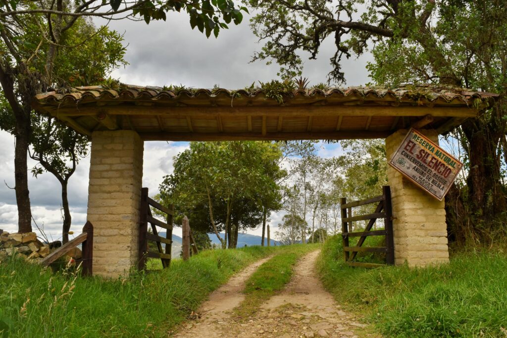 Fotografía del portón de entrada rústico de la Reserva Natural El Silencio en San Francisco, Cundinamarca. Se observa una estructura con columnas de ladrillo claro, techo de tejas cubierto de vegetación y un letrero de madera que dice 'Reserva Natural El Silencio - Conservemos el bosque andino de niebla', enmarcado por un paisaje verde de montaña.