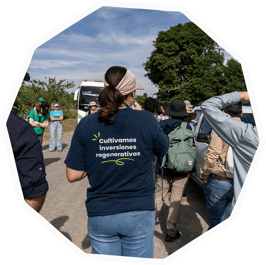 Vista posterior de una mujer con una camiseta azul que lleva impresa la frase 'Cultivamos inversiones regenerativas', rodeada de un grupo de personas en un día de campo y discusión sobre agroalimentación.