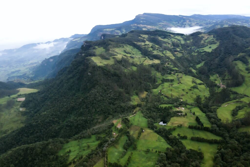 Toma aérea con dron de la Reserva Natural El Silencio en San Francisco, Cundinamarca. Muestra un paisaje montañoso de los Andes con pendientes pronunciadas cubiertas de densos parches de bosque andino y potreros verdes de ganadería silvopastoril bajo un cielo nublado
